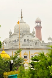 A majestic white building with intricate architectural details, featuring domes and spires. A red minaret stands prominently in the background, while lush green plants frame the foreground. A golden-yellow dome adds a pop of color, and an orange flag is prominently displayed.