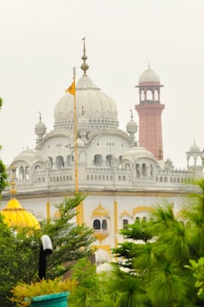 A majestic white building with intricate architectural details, featuring domes and spires. A red minaret stands prominently in the background, while lush green plants frame the foreground. A golden-yellow dome adds a pop of color, and an orange flag is prominently displayed.