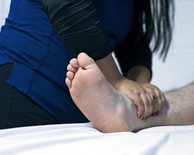 A person is receiving a foot massage, with hands gently gripping an ankle. The setting appears to be clinical or therapeutic, indicated by the professional attire of the masseur and the sterile environment.