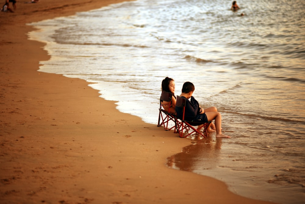 2 boys sitting on black folding chair on beach during daytime