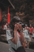 A person is taking a photo with a camera, surrounded by people in a lively outdoor setting. The individual is wearing a white shirt and a black watch, and a camera strap hangs from their neck. In the background, other people appear to be engaged in various activities, and there are red and natural earthy tones predominating in the scene.