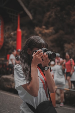 A person is taking a photo with a camera, surrounded by people in a lively outdoor setting. The individual is wearing a white shirt and a black watch, and a camera strap hangs from their neck. In the background, other people appear to be engaged in various activities, and there are red and natural earthy tones predominating in the scene.