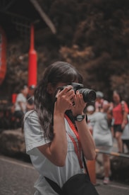 A person is taking a photo with a camera, surrounded by people in a lively outdoor setting. The individual is wearing a white shirt and a black watch, and a camera strap hangs from their neck. In the background, other people appear to be engaged in various activities, and there are red and natural earthy tones predominating in the scene.