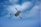 A drone is flying through a partly cloudy blue sky. The drone is prominently displayed, showing its four propellers and sleek design as it moves through the air.