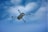 A drone is flying through a partly cloudy blue sky. The drone is prominently displayed, showing its four propellers and sleek design as it moves through the air.