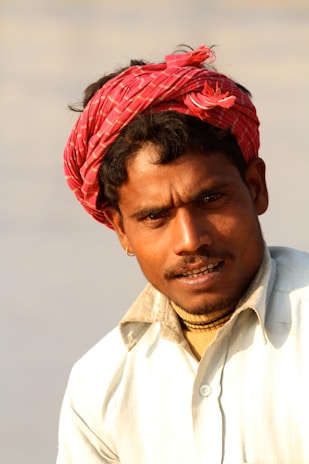A person wearing a bright red turban with a patterned design, coupled with a light-colored collared shirt. The lighting suggests a warm, possibly outdoor setting. The person's facial expression appears neutral, with a slight smile.