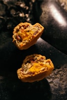 Two halves of a baked pastry filled with vibrant orange and yellow filling, containing visible seeds and nuts. The texture appears crumbly with a rich, moist interior. The pastries rest on a dark, stone-like surface with low lighting, creating a rustic and warm ambiance.