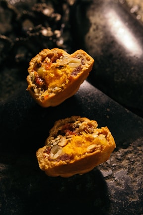 Two halves of a baked pastry filled with vibrant orange and yellow filling, containing visible seeds and nuts. The texture appears crumbly with a rich, moist interior. The pastries rest on a dark, stone-like surface with low lighting, creating a rustic and warm ambiance.