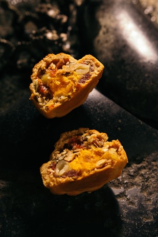 Two halves of a baked pastry filled with vibrant orange and yellow filling, containing visible seeds and nuts. The texture appears crumbly with a rich, moist interior. The pastries rest on a dark, stone-like surface with low lighting, creating a rustic and warm ambiance.
