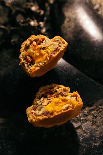 Two halves of a baked pastry filled with vibrant orange and yellow filling, containing visible seeds and nuts. The texture appears crumbly with a rich, moist interior. The pastries rest on a dark, stone-like surface with low lighting, creating a rustic and warm ambiance.