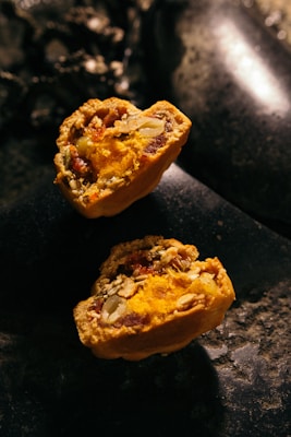 Two halves of a baked pastry filled with vibrant orange and yellow filling, containing visible seeds and nuts. The texture appears crumbly with a rich, moist interior. The pastries rest on a dark, stone-like surface with low lighting, creating a rustic and warm ambiance.