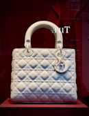 A white, quilted handbag with a structured design is displayed against a deep red background. The bag features metallic charms hanging from one of the handles, with circular rings attaching the handles to the body.