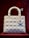 A white, quilted handbag with a structured design is displayed against a deep red background. The bag features metallic charms hanging from one of the handles, with circular rings attaching the handles to the body.