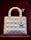 A white, quilted handbag with a structured design is displayed against a deep red background. The bag features metallic charms hanging from one of the handles, with circular rings attaching the handles to the body.