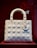 A white, quilted handbag with a structured design is displayed against a deep red background. The bag features metallic charms hanging from one of the handles, with circular rings attaching the handles to the body.
