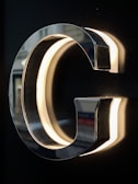 A chrome or metallic letter G is mounted on a dark background, with lighting on the inner edges creating a glowing effect. The reflective surface of the letter captures hints of the surroundings.