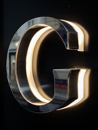 A chrome or metallic letter G is mounted on a dark background, with lighting on the inner edges creating a glowing effect. The reflective surface of the letter captures hints of the surroundings.