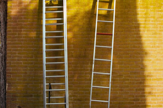Two aluminum ladders are propped against a yellow brick wall. Shadows from trees and the ladders are visible on the wall, creating an interesting play of light and shadow.