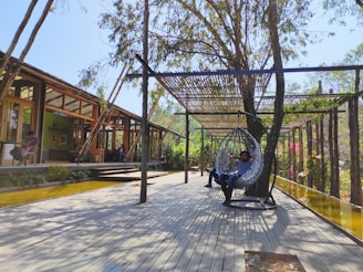 A modern outdoor patio area with wooden flooring and a wooden pergola providing some shade. Two people are relaxing; one in a hanging chair and another sitting at a table. The setting is surrounded by greenery and trees, creating a tranquil ambiance.