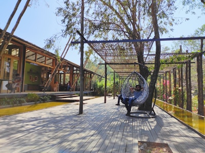 A modern outdoor patio area with wooden flooring and a wooden pergola providing some shade. Two people are relaxing; one in a hanging chair and another sitting at a table. The setting is surrounded by greenery and trees, creating a tranquil ambiance.
