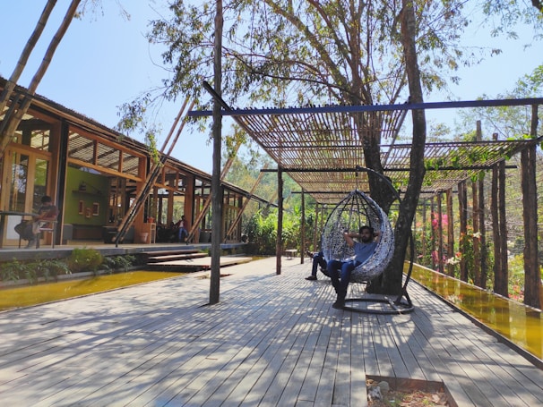 A modern outdoor patio area with wooden flooring and a wooden pergola providing some shade. Two people are relaxing; one in a hanging chair and another sitting at a table. The setting is surrounded by greenery and trees, creating a tranquil ambiance.