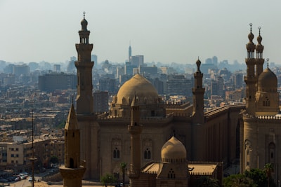 A cityscape featuring a large mosque with majestic domes and tall minarets, surrounded by numerous buildings of varying heights. The architecture displays intricate Islamic design elements, standing prominently against a backdrop of a sprawling urban skyline.