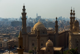 A cityscape featuring a large mosque with majestic domes and tall minarets, surrounded by numerous buildings of varying heights. The architecture displays intricate Islamic design elements, standing prominently against a backdrop of a sprawling urban skyline.