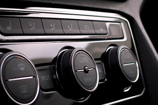 Close-up of a car's climate control panel featuring two large knobs with embedded display screens and smaller buttons for various functions. The surfaces are glossy with metallic and black elements.
