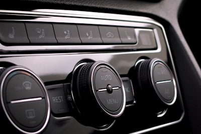 Close-up of a car's climate control panel featuring two large knobs with embedded display screens and smaller buttons for various functions. The surfaces are glossy with metallic and black elements.