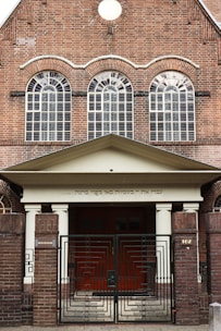 The image features a brick building with three large arched windows. Ornate metal gates stand at the entrance. There is a Hebrew inscription just above the entrance, supported by two columns.