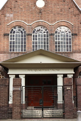 The image features a brick building with three large arched windows. Ornate metal gates stand at the entrance. There is a Hebrew inscription just above the entrance, supported by two columns.