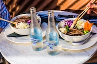 Two bowls of food are served on a round marble table, each bowl containing a mix of colorful vegetables including shredded cabbage, sliced carrots, and mushrooms, accompanied by a bottle of sparkling water. A hand is using chopsticks to eat from one of the bowls.