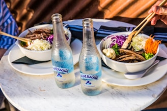 Two bowls of food are served on a round marble table, each bowl containing a mix of colorful vegetables including shredded cabbage, sliced carrots, and mushrooms, accompanied by a bottle of sparkling water. A hand is using chopsticks to eat from one of the bowls.