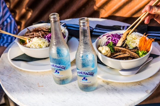 Two bowls of food are served on a round marble table, each bowl containing a mix of colorful vegetables including shredded cabbage, sliced carrots, and mushrooms, accompanied by a bottle of sparkling water. A hand is using chopsticks to eat from one of the bowls.