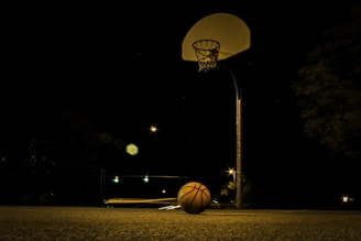 A dimly lit outdoor basketball court at night with a basketball resting on the ground beneath the hoop. The court is surrounded by darkness, with faint lights and star-like points in the distance creating a moody atmosphere.