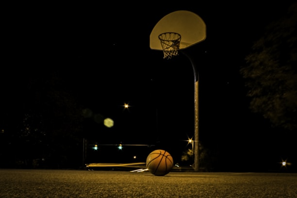 A dimly lit outdoor basketball court at night with a basketball resting on the ground beneath the hoop. The court is surrounded by darkness, with faint lights and star-like points in the distance creating a moody atmosphere.