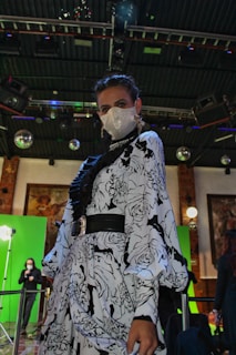 A person is wearing an ornate black and white floral patterned dress with a sparkly face mask and elaborate earrings, standing in an indoor setting under a high ceiling. The background features bright lights, disco balls, and green screens, contributing to a sophisticated event or photoshoot atmosphere.