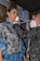 A person wearing a patterned garment is being assisted by another individual wearing a camouflage-patterned jacket and a protective mask. They appear to be in a dimly lit room with some colorful artwork on the walls in the background.