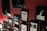 A red textured wall displaying an assortment of framed posters and photographs. The frames vary in size and orientation, creating a collage-like effect. Some posters feature text, while others show images of landscapes and abstract designs.