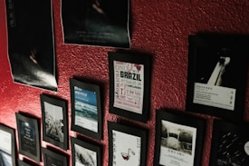 A red textured wall displaying an assortment of framed posters and photographs. The frames vary in size and orientation, creating a collage-like effect. Some posters feature text, while others show images of landscapes and abstract designs.