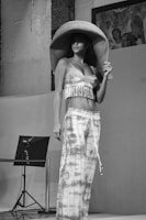 A person wearing a large, wide-brimmed hat, a fringed top, and loose patterned pants stands indoors. The setting includes a painted artwork on the wall and a lighting or sound setup in the background.