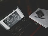 A dimly lit study setup with a laptop displaying a diagrammatic sketch with arrows and labels. Beside the laptop is a notebook filled with handwritten notes, a calculator, and a pen resting on top.