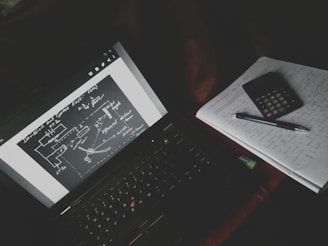 A dimly lit study setup with a laptop displaying a diagrammatic sketch with arrows and labels. Beside the laptop is a notebook filled with handwritten notes, a calculator, and a pen resting on top.