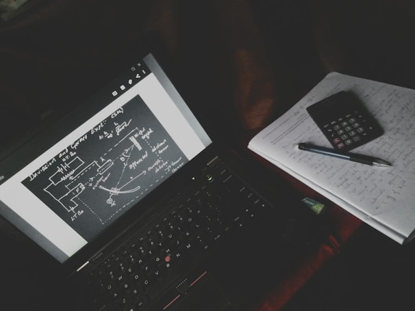 A dimly lit study setup with a laptop displaying a diagrammatic sketch with arrows and labels. Beside the laptop is a notebook filled with handwritten notes, a calculator, and a pen resting on top.