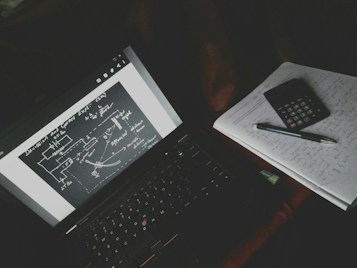 A dimly lit study setup with a laptop displaying a diagrammatic sketch with arrows and labels. Beside the laptop is a notebook filled with handwritten notes, a calculator, and a pen resting on top.