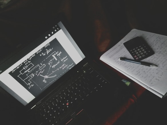 A dimly lit study setup with a laptop displaying a diagrammatic sketch with arrows and labels. Beside the laptop is a notebook filled with handwritten notes, a calculator, and a pen resting on top.