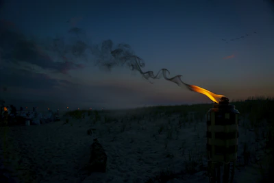 A cozy night beach party scene with tiki torches glowing softly, Japanese lanterns hanging overhead, and a colorful LGBTQIA flag gently waving in the breeze.