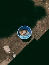 A blue plastic bucket containing trash, including cardboard and blue fabric, placed on a rough, dark surface. There are shadows casting over parts of the ground, creating a contrast, and a small portion of a shoe partially visible at the bottom.