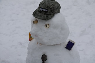 A snowman is adorned with various accessories including a camouflage Supreme cap, a small carton of juice, sunglasses, and a smartphone. The snowman's facial features are creatively made from various objects.