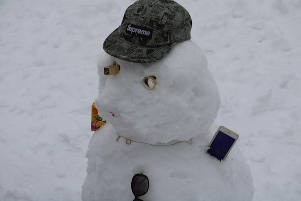A snowman is adorned with various accessories including a camouflage Supreme cap, a small carton of juice, sunglasses, and a smartphone. The snowman's facial features are creatively made from various objects.